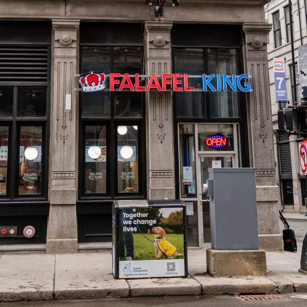 Falafel King restaurant storefront with a red and blue sign, an "OPEN" neon sign, and a street-side advertisement.