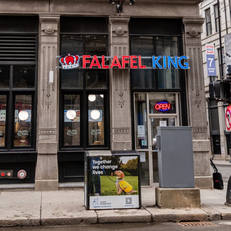 Falafel King restaurant storefront with a red and blue sign, an "OPEN" neon sign, and a street-side advertisement.