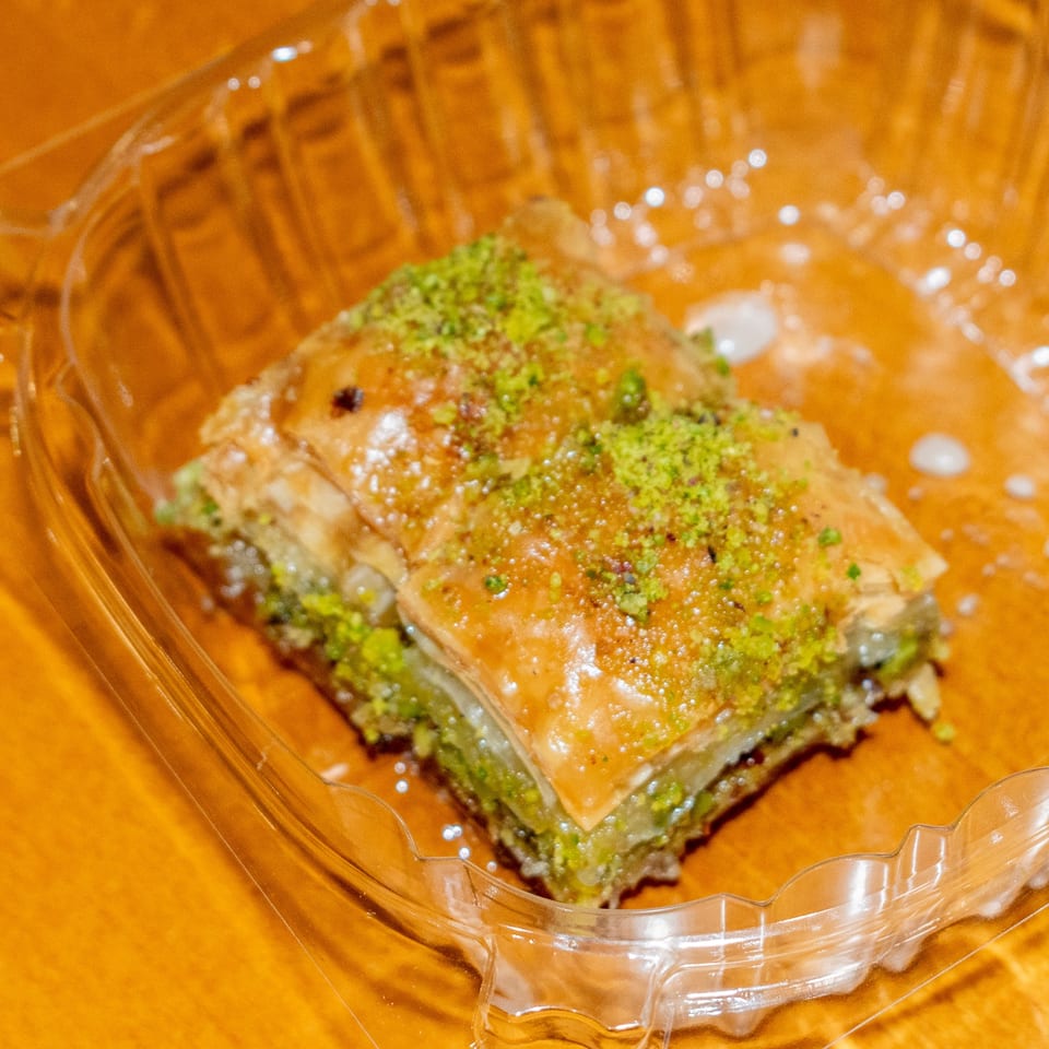 A piece of baklava with green pistachio crumbles and honey glaze in a clear plastic container.