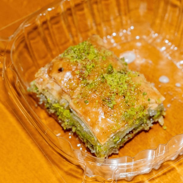 A piece of baklava with green pistachio crumbles and a honey glaze in a plastic container.