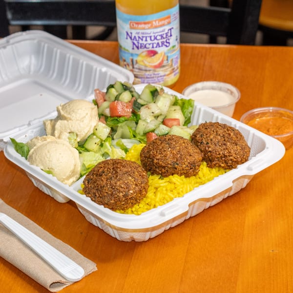 Falafel King takeout container with falafel, yellow rice, hummus, salad, and a drink on a wooden table.